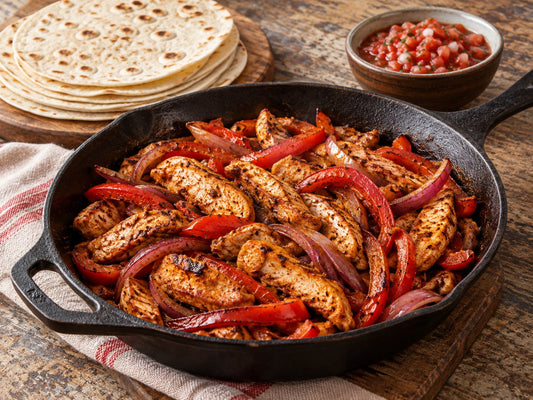 Cooked chicken breast strips, red pepper and red onion strips coated with smoky reddish spices in a plain iron skillet; plain tortillas and small bowl of tomato salsa alongside, generated illustration